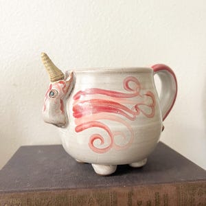 May include: A whimsical ceramic unicorn mug. The mug is off-white with pink and red swirled designs. It features a unicorn horn, a handle with a red accent, and three small feet. The mug is a unique piece of drinkware.