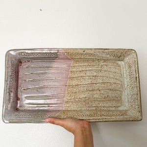 May include: Rectangular ceramic tray with a two-tone design. One side is light pink with horizontal grooves, the other is tan with textured lines. The tray has a speckled, textured border.
