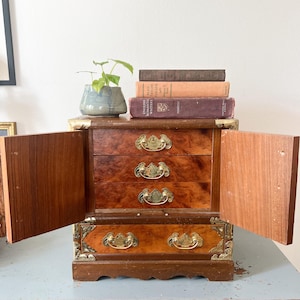 May include: A small, antique wooden jewelry box with three drawers and brass hardware. The box has two doors that are open, revealing the interior. A small potted plant and several books are on top of the box.