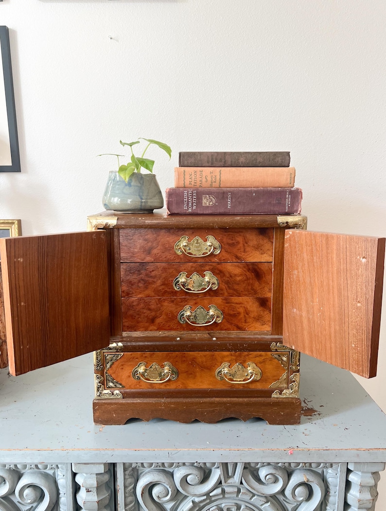 May include: A small, antique wooden jewelry box with three drawers and brass hardware. The box has two doors that are open, revealing the interior. A small potted plant and several books are on top of the box.
