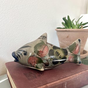 May include: A ceramic fish figurine with a gray base color, adorned with floral patterns in shades of green, pink, and black. The fish has black stripes and a blue mouth. It rests on a maroon book.