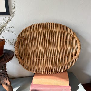 May include: An oval-shaped woven wicker tray featuring a natural light brown finish and two integrated side handles. The tray is constructed with vertical reed slats and a braided border, showcasing a textured, open-weave design throughout.