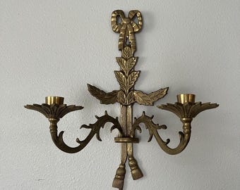 May include: A gold-toned brass wall sconce with two candle holders. The sconce features a decorative bow at the top and ornate leaf designs on the arms.