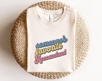 Puede incluir: Una camiseta blanca con el texto "someone's favorite accountant" en una fuente retro con colores azul, amarillo, rosa y naranja.