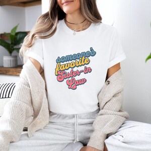 Könnte beinhalten: Weißes T-Shirt mit dem Text "someone's favorite Sister-in-Law" in einem Retro-Schriftstil. Der Text ist in Blau, Gelb und Rosa gehalten. Das Shirt hat einen Rundhalsausschnitt und kurze Ärmel.