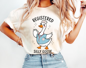 Puede incluir: Una camiseta blanca con un dibujo animado de un ganso vestido de enfermera, con un estetoscopio. El texto "Registered Silly Goose" está impreso en la camiseta.