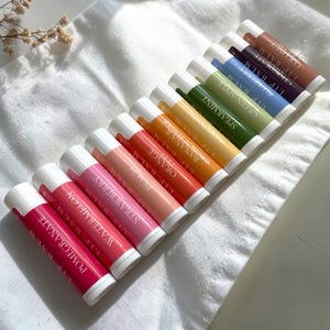 Op de afbeelding: Een assortiment kleurrijke lippenbalsems, gerangschikt op een witte doek. De lippenbalsems zijn in verschillende tinten, waaronder roze, oranje, groen en blauw, met labels die smaken aangeven zoals granaatappel, watermeloen en aardbei.