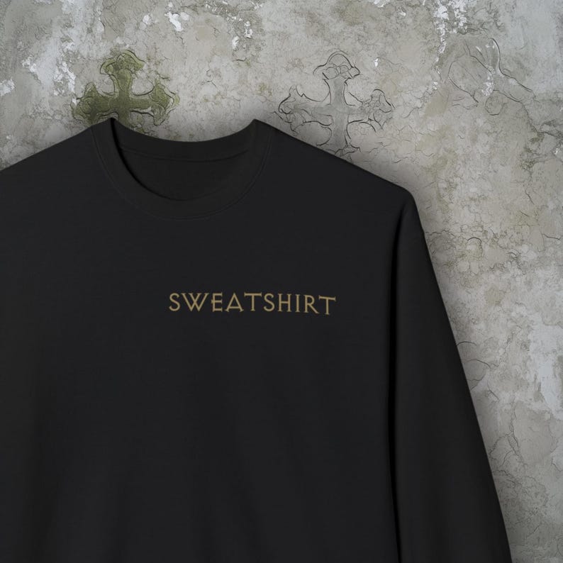 Puede incluir: Sudadera negra con la palabra "SWEATSHIRT" en letras doradas. La sudadera es de material suave y tiene cuello redondo. El fondo es una pared gris texturizada con un dise&ntilde;o de cruz.