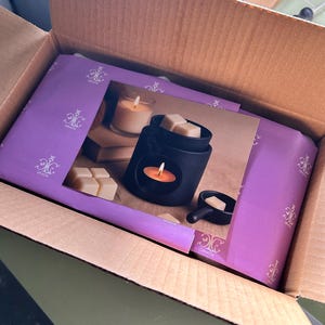 May include: A black wax warmer with a candle and wax cubes, packaged in a cardboard box. The warmer is displayed on a purple box with a decorative design. A Fire TV Stick box is visible in the background.