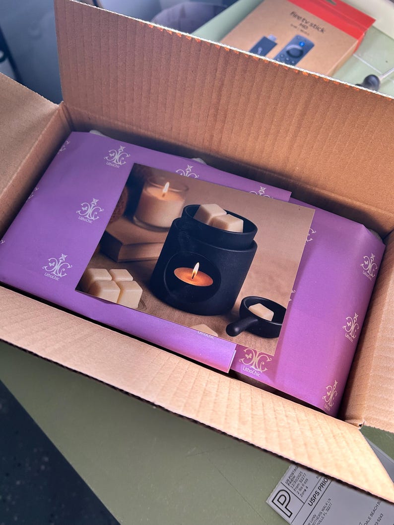 May include: A black wax warmer with a candle and wax cubes, packaged in a cardboard box. The warmer is displayed on a purple box with a decorative design. A Fire TV Stick box is visible in the background.