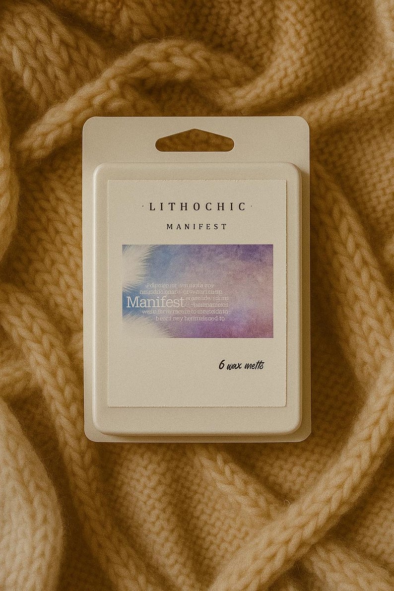 May include: A package of six wax melts, labeled "LITHOCHIC MANIFEST." The label features a watercolor design in shades of purple and blue, with the word "Manifest" in a stylized font. The package is white and sits on a beige knitted surface.