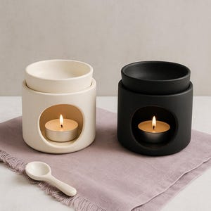 May include: Two ceramic oil warmers, one cream and one black, each with a tea light candle inside. A small white spoon rests on a lavender-colored cloth. The warmers have a circular opening to hold the candle.