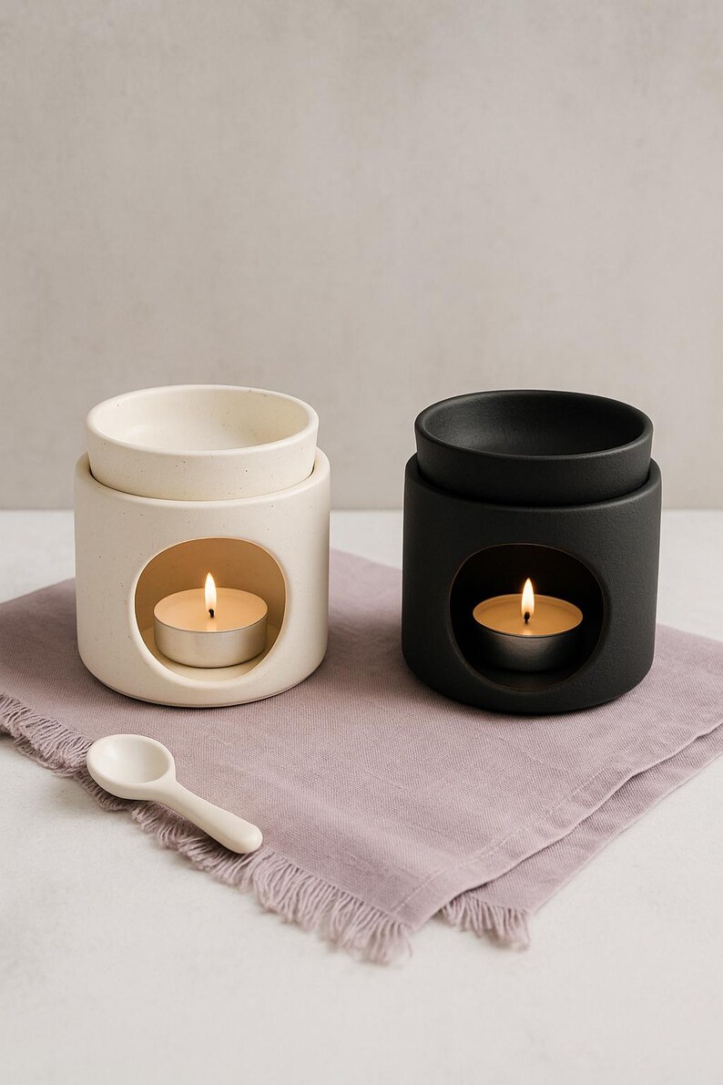May include: Two ceramic oil warmers, one cream and one black, each with a tea light candle inside. A small white spoon rests on a lavender-colored cloth. The warmers have a circular opening to hold the candle.