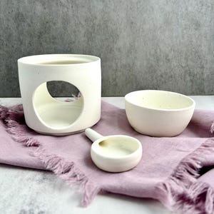 May include: A white ceramic oil diffuser with a circular opening, a small bowl, and a spoon-shaped dish, all arranged on a lavender-colored cloth. The objects are set against a gray background, creating a minimalist aesthetic.