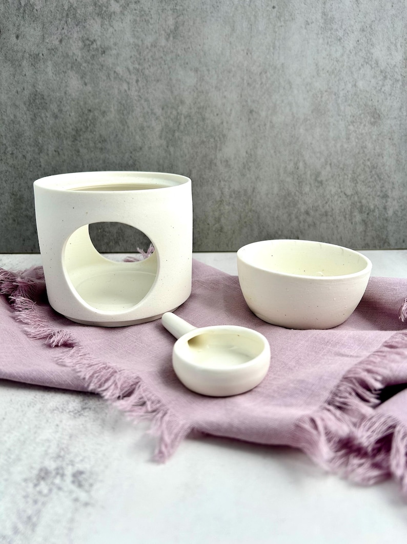 May include: A white ceramic oil diffuser with a circular opening, a small bowl, and a spoon-shaped dish, all arranged on a lavender-colored cloth. The objects are set against a gray background, creating a minimalist aesthetic.