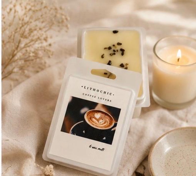 May include: A package of Lithochic Coffee Lovers wax melts with a coffee art image. The wax melts are light yellow with dark specks. A lit candle in a glass and a small ceramic dish are also in the image.
