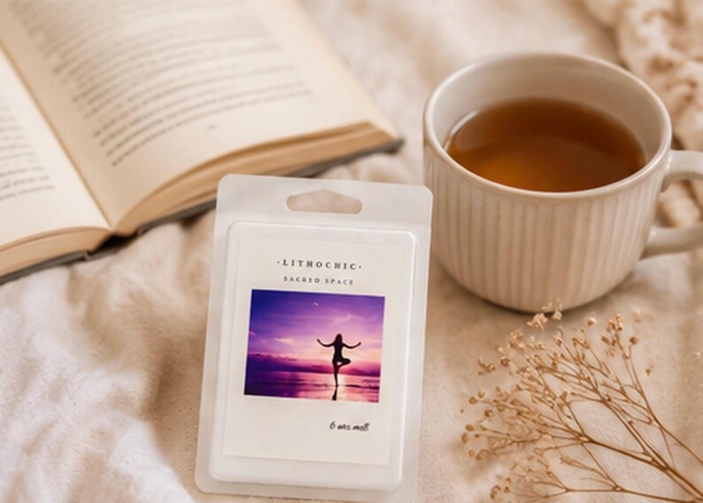 May include: A white wax melt package with the text "LITHOCHIC - SACRED SPACE" and an image of a person doing yoga at sunset. A cup of tea, an open book, and dried flowers are also in the image.