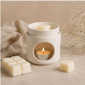 May include: A cream-colored ceramic wax warmer with a lit tea light inside. The warmer has a round opening and a top dish holding a wax melt. A tray of wax melts and a single melt are also visible, suggesting a home fragrance theme.