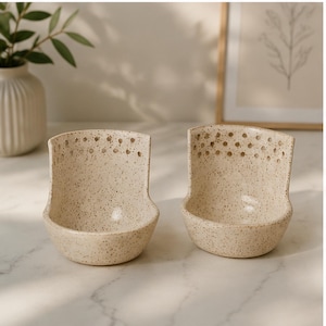 May include: Two speckled ceramic spoon rests. Each rest has a curved, open bowl shape with a flat base and a series of small holes near the top. The color is a light beige with darker speckles, and they sit on a white surface.