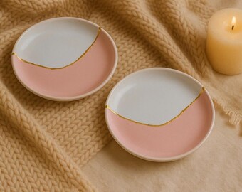 Puede incluir: Dos platos pequeños y redondos de cerámica, con un diseño blanco y rosa, separados por una línea dorada. Una vela encendida está cerca. Los platos están sobre una superficie beige tejida.