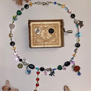 May include: A silver chain necklace with a variety of colorful beads, charms, and pendants, including hearts, butterflies, and a dragonfly. The necklace is displayed on a white background with a wooden music box.