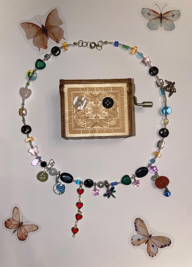 May include: A silver chain necklace with a variety of colorful beads, charms, and pendants, including hearts, butterflies, and a dragonfly. The necklace is displayed on a white background with a wooden music box.