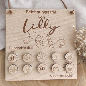 Könnte beinhalten: Belohnungstafel aus Holz mit dem Namen "Lilly" und einer Schildkröten-Illustration. Enthält Holz-Token mit Meereskreaturen-Designs. Der Text "Du schaffst das!" und "Super gemacht!" sind ebenfalls sichtbar.
