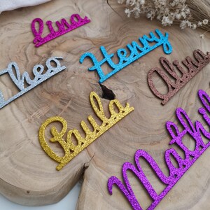 Könnte beinhalten: Glitzernde Namensausstanzungen in verschiedenen Farben, darunter Rosa, Blau, Gold, Silber, Braun und Lila, auf einer Holzoberfläche angeordnet. Namen wie Luna, Henry, Theo, Paula und andere sind zu sehen. Ideal für personalisierte Geschenke.