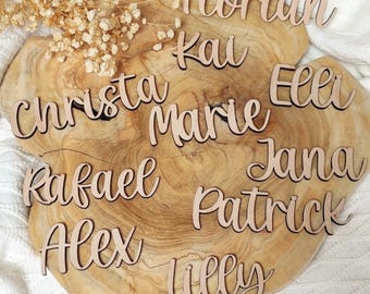Puede incluir: Tarjetas de nombre de madera con los nombres: Christa, Florian, Kai, Marie, Elli, Rafael, Alex, Jana, Patrick y Lilly. Las tarjetas de nombre están sobre una superficie de madera.