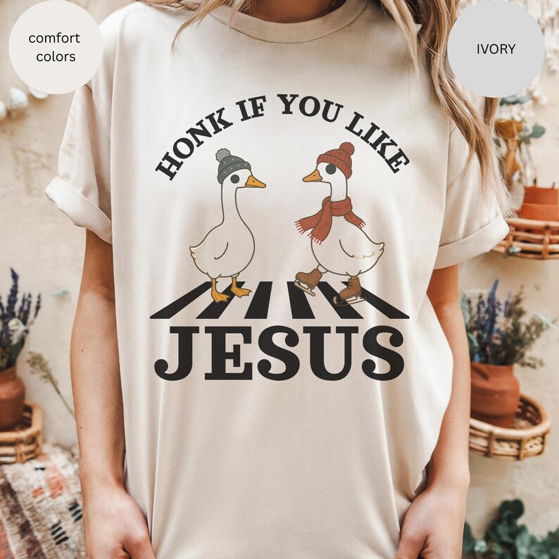 May include: Ivory-colored t-shirt with the text "HONK IF YOU LIKE JESUS." The graphic features two cartoon geese wearing hats and scarves, crossing a crosswalk. The shirt has comfort colors.