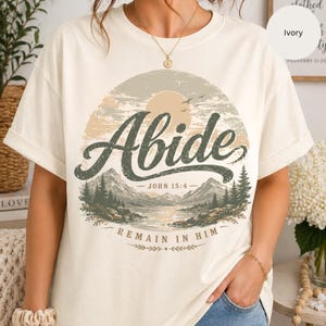 May include: Ivory-colored t-shirt featuring a scenic landscape design. The design includes the word "Abide" in a stylized font, with mountains, a lake, and the text "John 15:4" and "Remain in Him."