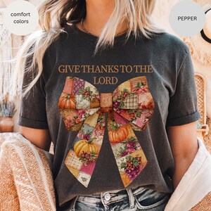May include: Dark gray t-shirt with the text "GIVE THANKS TO THE LORD" above a large bow design. The bow features a patchwork pattern with pumpkins, grapes, and floral accents in shades of orange, yellow, and purple. The shirt is labeled "comfort colors" and "pepper".