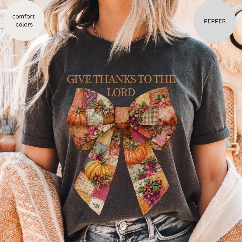 May include: Dark gray t-shirt with the text "GIVE THANKS TO THE LORD" above a large bow design. The bow features a patchwork pattern with pumpkins, grapes, and floral accents in shades of orange, yellow, and purple. The shirt is labeled "comfort colors" and "pepper".