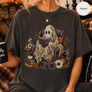 Peut inclure: Un t-shirt gris anthracite orné d'un motif représentant un fantôme assis dans un fauteuil violet en train de lire un livre. Le fantôme est entouré de fleurs, de livres empilés, d'une citrouille, d'une tasse à cœur et d'une lanterne aux tons violets.