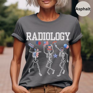 Może przedstawiać: Szary T-shirt z nadrukiem "RADIOLOGY" w kolorze białym. Na koszulce znajduje się grafika przedstawiająca trzy szkielety w czerwono-biało-niebieskich patriotycznych dodatkach, w tym kapeluszu cylindrycznym, okularach przeciwsłonecznych i fladze.