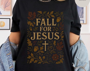 May include: Black t-shirt with a fall-themed design. The design features the words "FALL FOR JESUS" in a gold color, surrounded by autumn leaves, acorns, and a cross. The shirt is a classic crew neck.