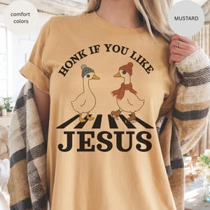 May include: Mustard-colored t-shirt with the text "HONK IF YOU LIKE JESUS." The graphic features two cartoon geese on a crosswalk, one wearing a hat and the other a scarf and ice skates. The shirt is a comfort color.