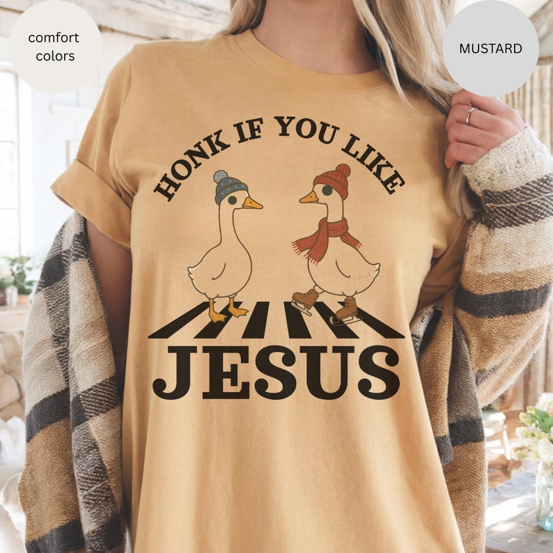 May include: Mustard-colored t-shirt with the text "HONK IF YOU LIKE JESUS." The graphic features two cartoon geese on a crosswalk, one wearing a hat and the other a scarf and ice skates. The shirt is a comfort color.