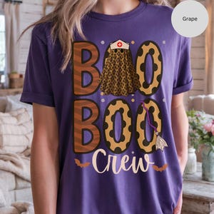 May include: A purple t-shirt with a Halloween-themed graphic. The design features the words "BOO BOO CREW" in brown and gold lettering, with a leopard print nurse's outfit and a witch's broom. Two small bats are at the bottom.