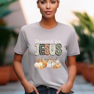 Op de afbeelding: Een grijs T-shirt met de tekst "Thankful for JESUS" in een kleurrijk, decoratief lettertype. De tekst wordt omringd door een herfstthema-ontwerp met pompoenen, bladeren en een lantaarn.