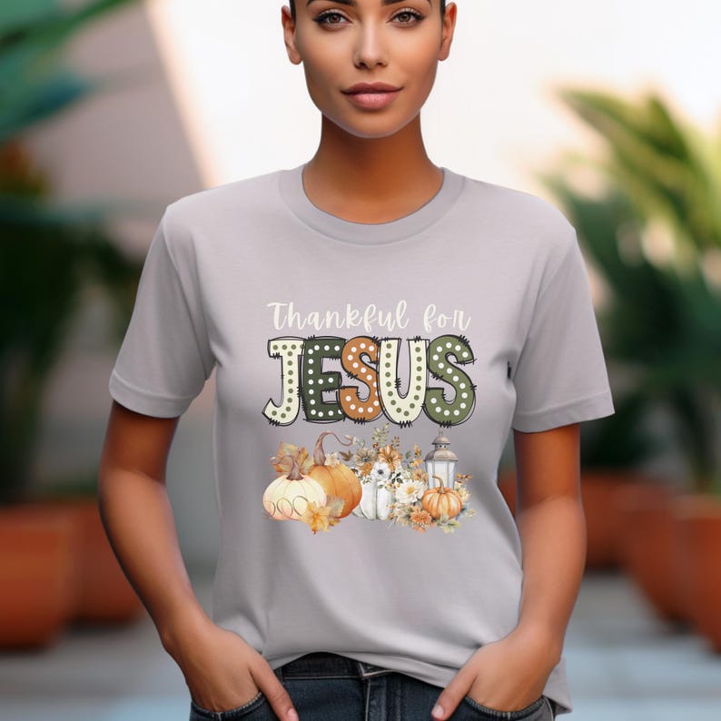 Op de afbeelding: Een grijs T-shirt met de tekst "Thankful for JESUS" in een kleurrijk, decoratief lettertype. De tekst wordt omringd door een herfstthema-ontwerp met pompoenen, bladeren en een lantaarn.