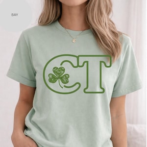 May include: A pale green t-shirt with a green graphic. The design shows the letters "CT" with a shamrock in the "C". The word "BAY" is in the upper left corner of the shirt.
