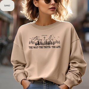 May include: A tan sweatshirt with a mountain scene and a cross. The text "JOHN 14:6" and "THE WAY-THE TRUTH-THE LIFE" are printed on the sweatshirt.