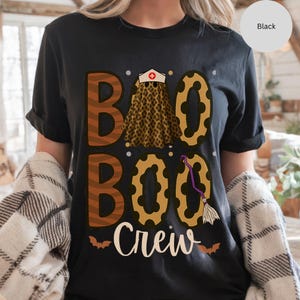 May include: Black t-shirt with the words "Boo Boo Crew" in white script. The word "Boo" is designed with a leopard print skirt and a nurse's cap. A purple broom and bats complete the Halloween theme.