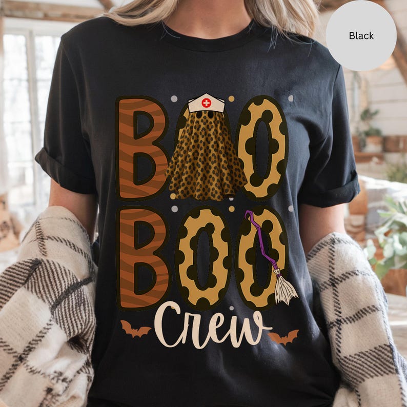 May include: Black t-shirt with the words "Boo Boo Crew" in white script. The word "Boo" is designed with a leopard print skirt and a nurse's cap. A purple broom and bats complete the Halloween theme.