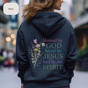 Pu&ograve; includere: Una felpa con cappuccio blu navy con un design floreale e il testo "blessed by GOD loved by JESUS lead by the SPIRIT" stampato sul retro.