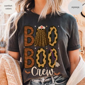 May include: Dark gray t-shirt with the words "BOO BOO Crew" in brown and gold lettering. The design includes a nurse's cap, a leopard print skirt, a witch's broom, and bats, perfect for Halloween.