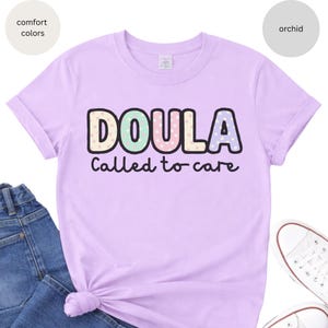 May include: A lilac t-shirt featuring the word "DOULA" in colourful polka-dot lettering and the phrase "Called to care" in black. The shirt is styled with blue jeans and white trainers.