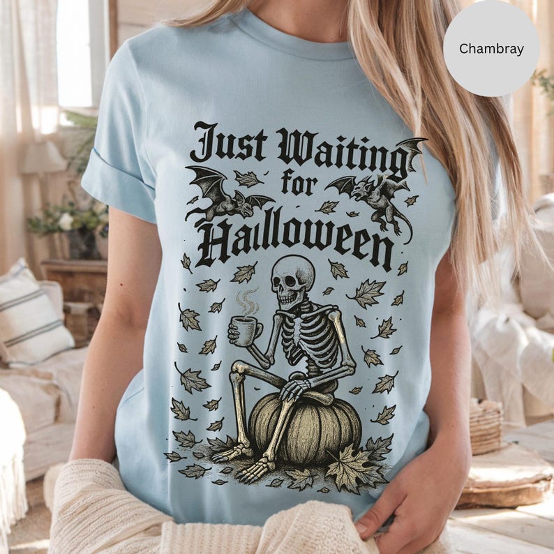 May include: Light blue t-shirt with the text "Just Waiting for Halloween". The graphic features a skeleton sitting on a pumpkin, holding a mug, with bats and leaves. The shirt is a Chambray color.