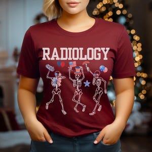 Może przedstawiać: Bordowy t-shirt z napisem "RADIOLOGY" w białych literach. Trzy szkielety noszą czapki imprezowe i trzymają amerykańskie flagi i balony.